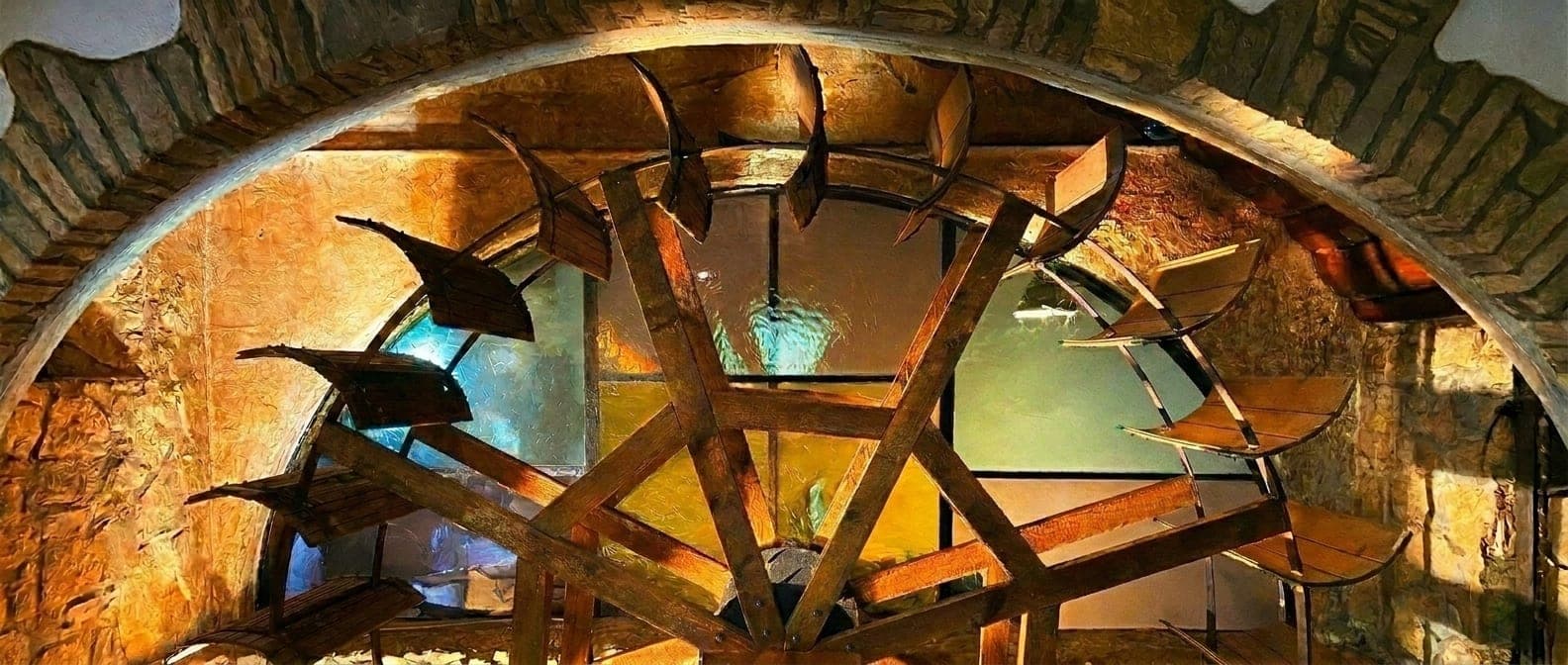 Großes Kunstglas-Fenster mit Backstein-Bogen, dahinter das historische Mühlrad der KnochenMühle